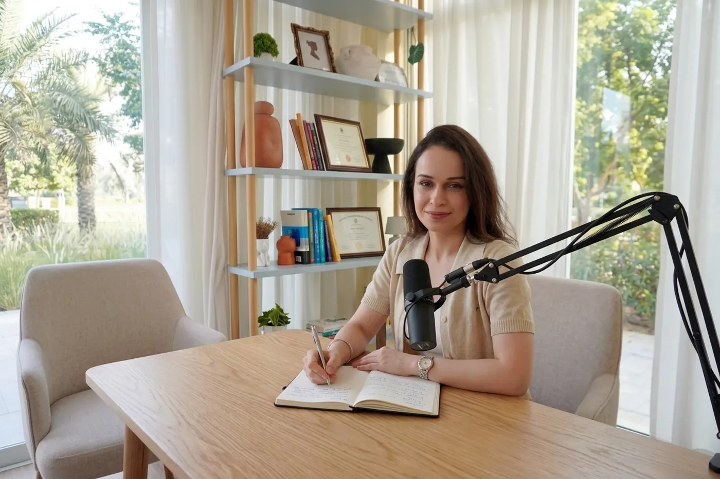 Komel Kaur at her desk with a podcast microphone, writing in a notebook, framed psychology credentials behind her